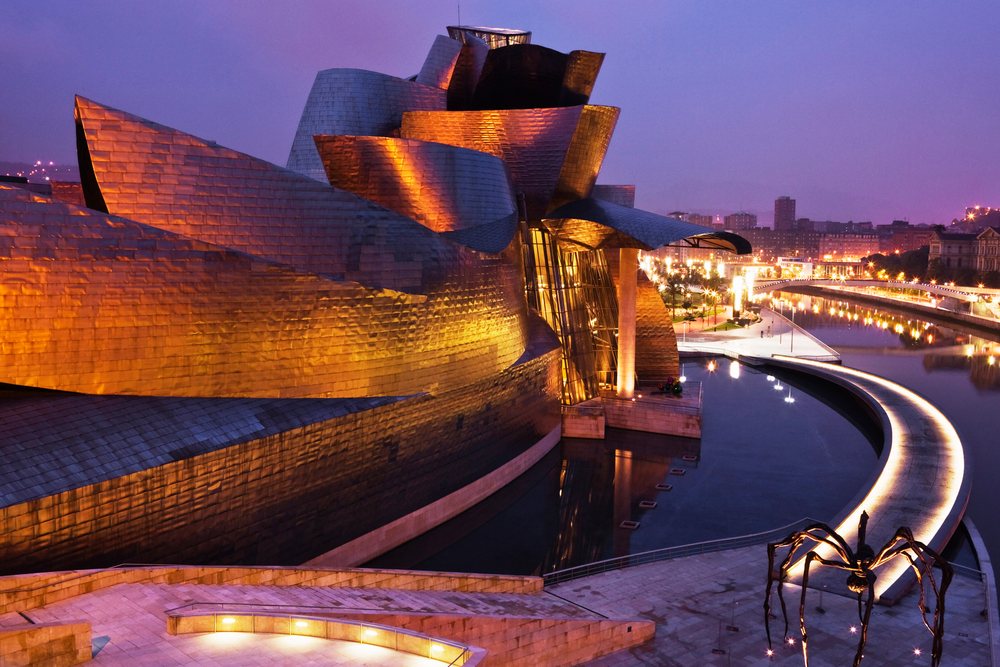 The Guggenheim Museum Bilbao illuminated at dusk by the river with its distinctive modern architecture and nearby large spider sculpture on the plaza, Spain.