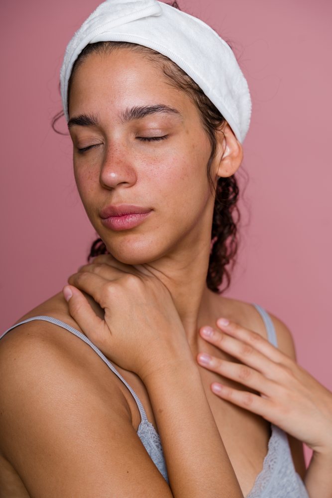 Woman with closed eyes wearing a white headband and grey strap top touching her shoulder against a pink background in a beauty shoot, Spain.
