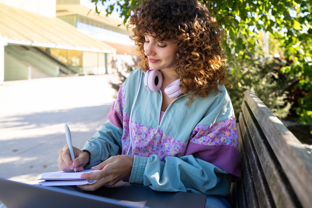 Woman with curly hair sitting on a bench outdoors in a park, writing in a notebook with a laptop and purple headphones nearby, Spain.