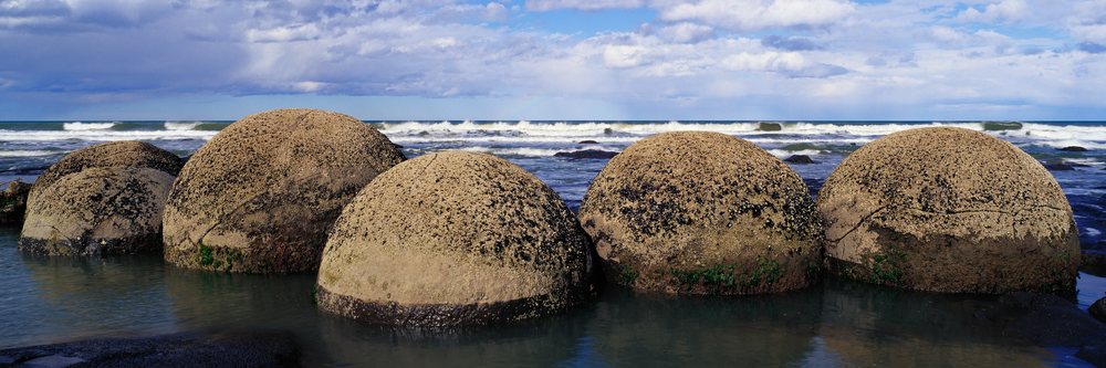 Large spherical Moeraki boulders with barnacles and moss arranged near water's edge on a beach with ocean waves under a cloudy sky.