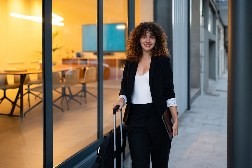 Young business woman standing outside a modern office building in Madrid holding a tablet and rolling suitcase.