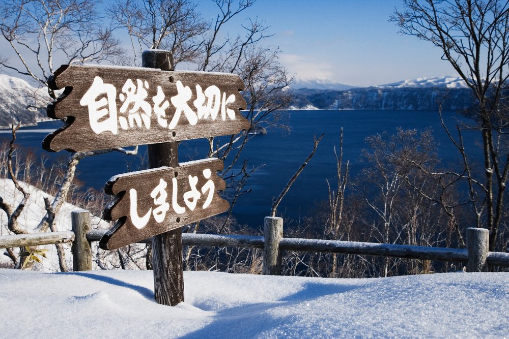 Wooden sign with Japanese text stands in snow Park, Hokkaido, with winter trees and snow-capped mountains visible, Japan, Asia.