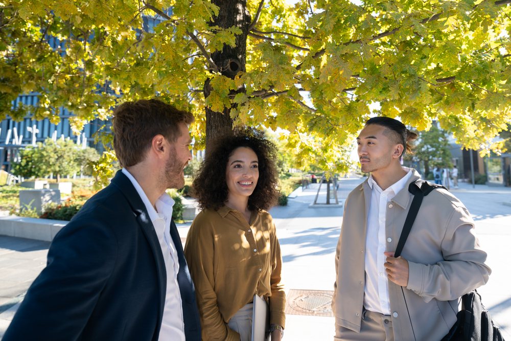 Three young adults engaged in conversation outdoors under a tree with green and yellow leaves in an urban setting, Madrid, Spain.