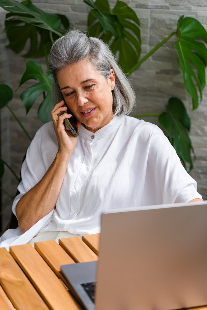 Woman talking on phone while using laptop at wooden table indoors with large green plants and stone wall background, Spain.