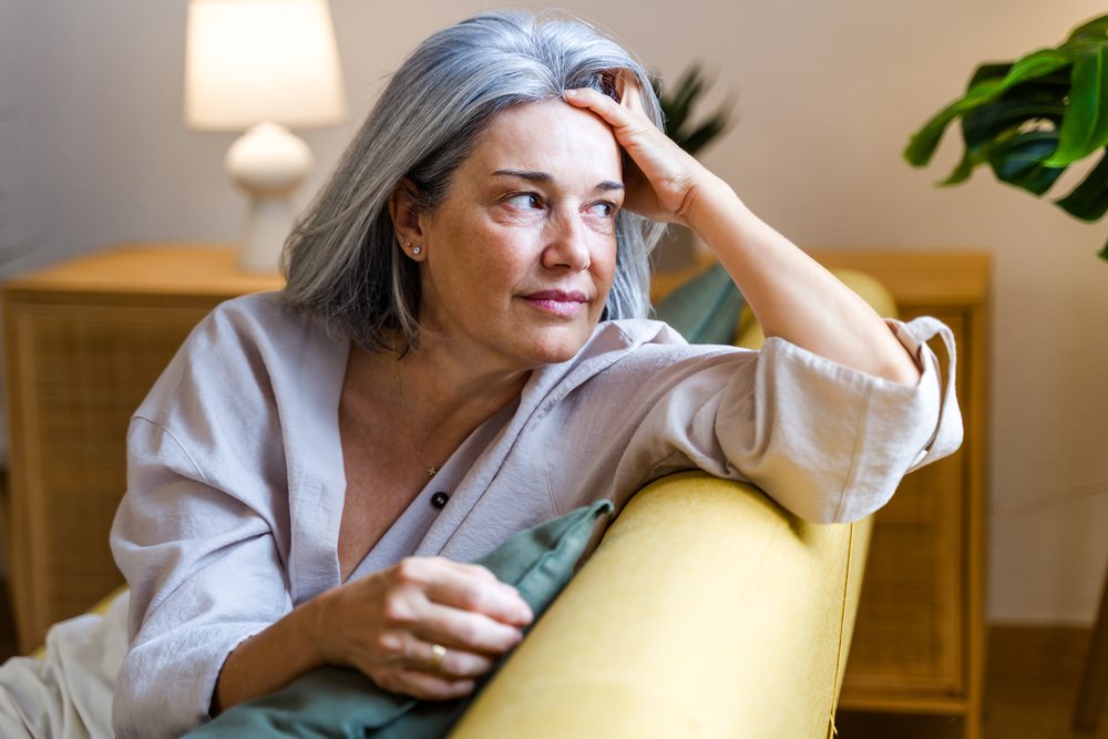Middle-aged woman with gray hair sitting thoughtfully on a yellow couch in a modern home interior, Spain.