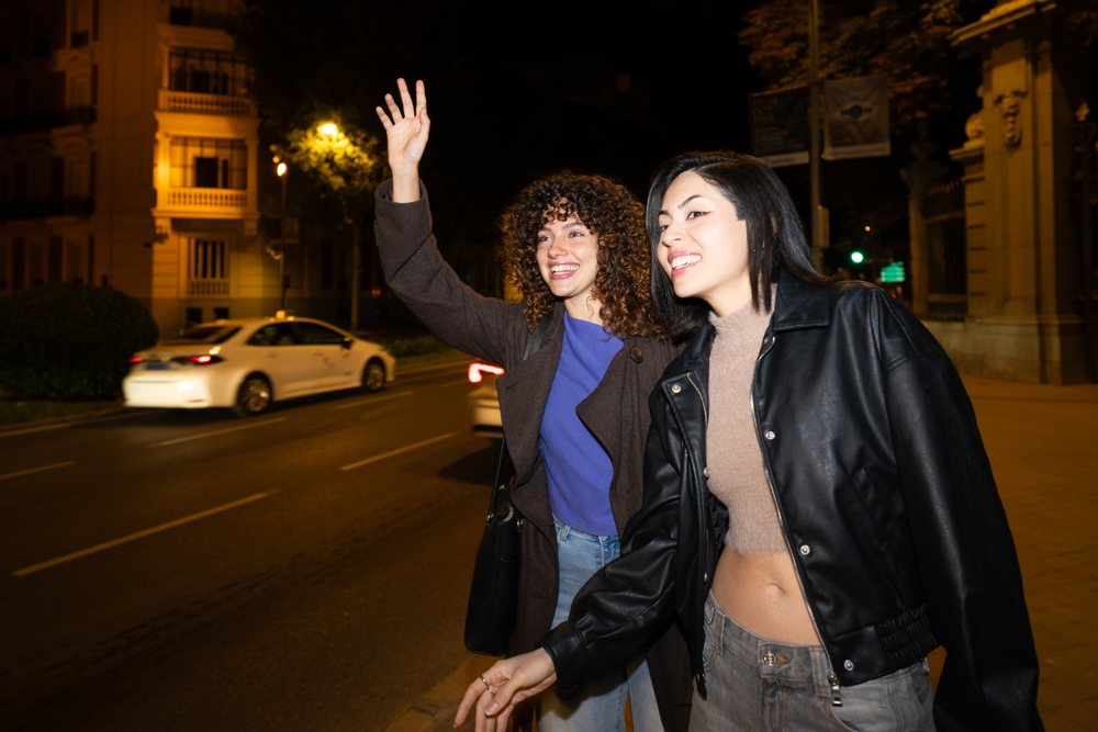 Two women smiling and walking outside at night on a city street, while one waves to hail a taxi, Madrid, Spain.