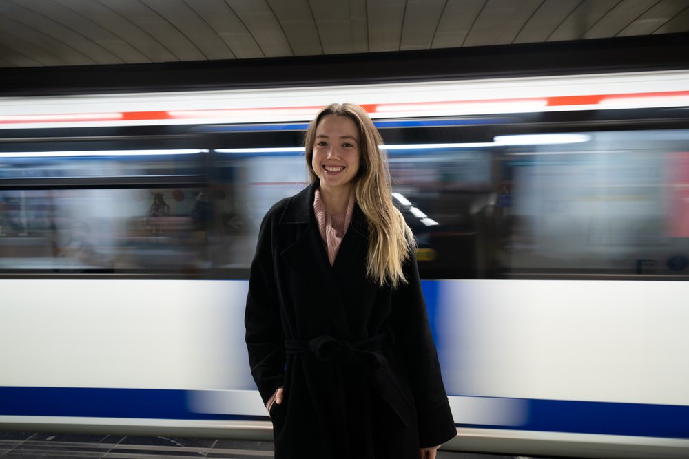 Woman in a black coat smiles on a subway platform with a moving train behind her, Christmas market, Madrid, Spain.
