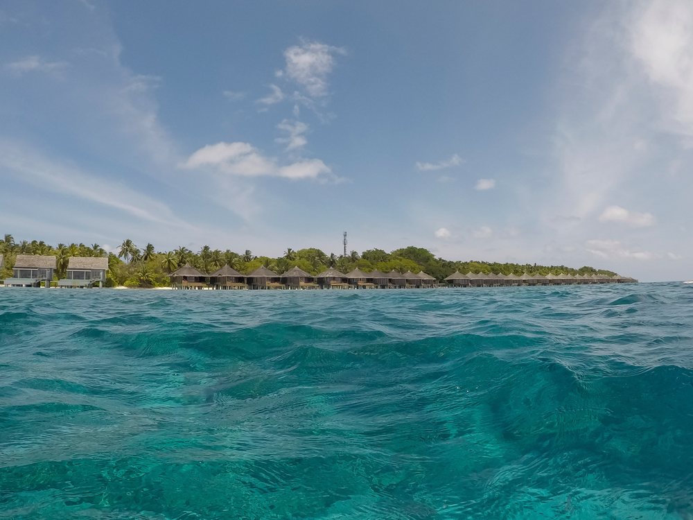 Clear turquoise waters in front of a tropical island with overwater bungalows under a blue sky with scattered clouds, Maldives.