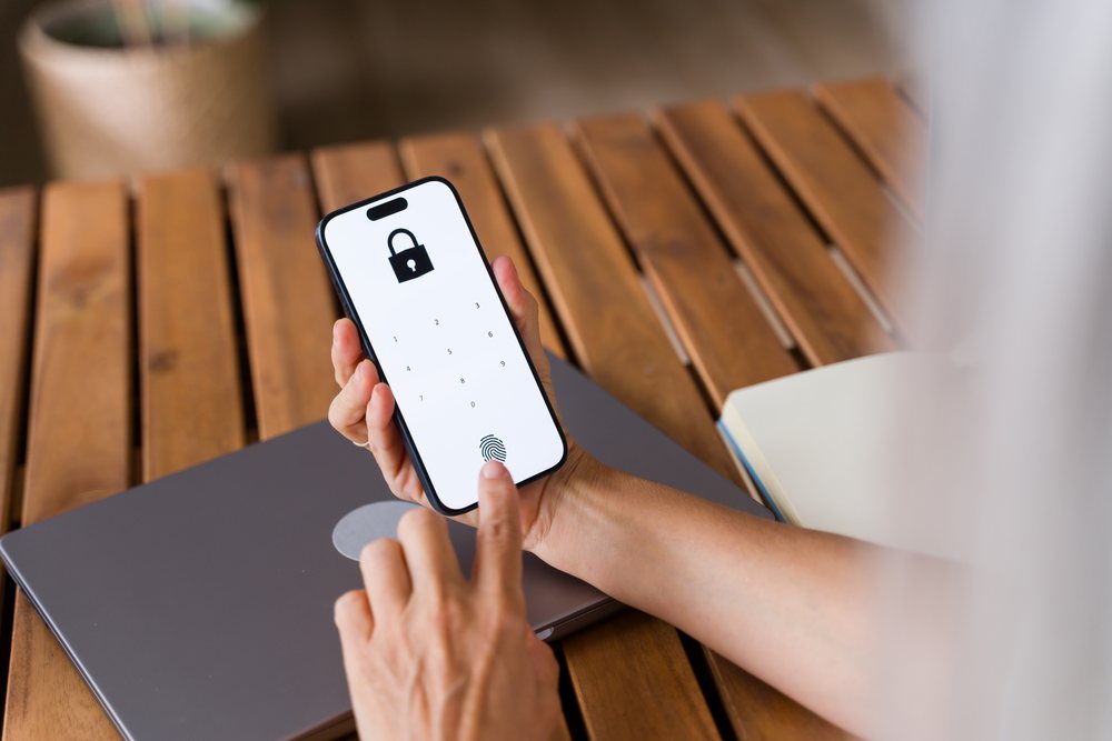 Person unlocking a smartphone with a fingerprint on its digital lock screen, with a laptop and notebook on a wooden table, Spain.