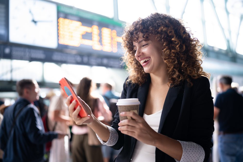 Woman with curly hair smiles holding a paper coffee cup and smartphone at a busy indoor train station waiting area, Spain.