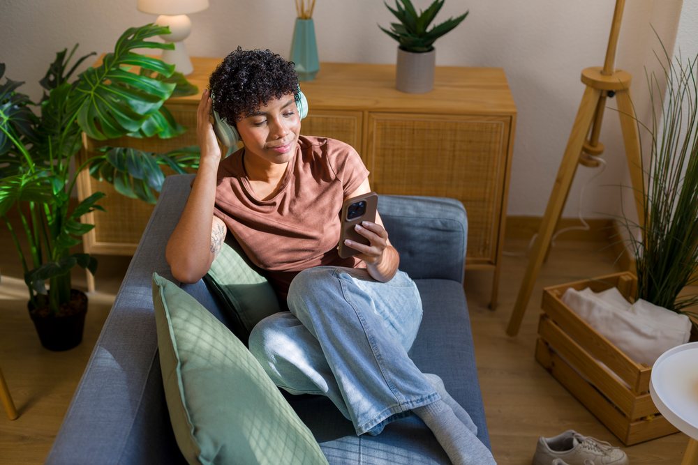 Young woman relaxing on a sofa while listening to music through headphones and using a smartphone at home.