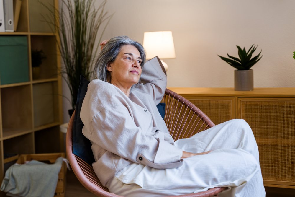 Woman with gray hair relaxing thoughtfully seated in a modern armchair in a stylish indoor home interior with lamp and houseplant nearby, Spain.