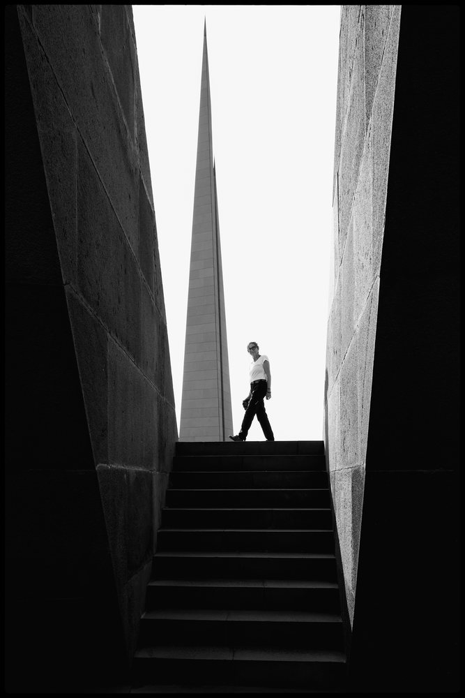 Person walking on stairs near the tall vertical spire of the Tsitsernakaberd memorial, in a black and white photograph, Yerevan, Armenia.