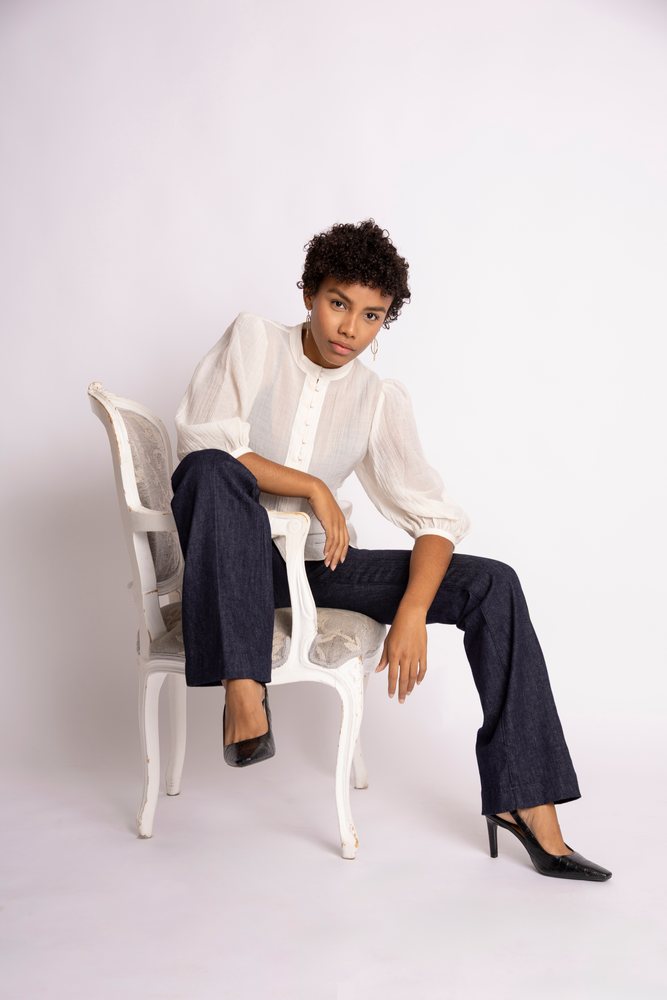 Woman seated on an antique white chair wearing a white puff sleeve blouse and dark trousers in a studio setting, fashion, young culture.