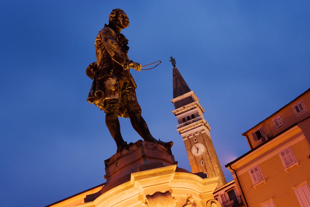 Statue of Giuseppe Tartini holding a violin bow in an urban square at dusk with a clock tower and historic buildings nearby, Piran, Slovenia.