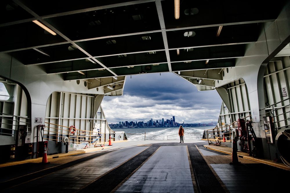 A person stands on the vehicle deck of a ferry overlooking an urban skyline under a cloudy sky, USA.