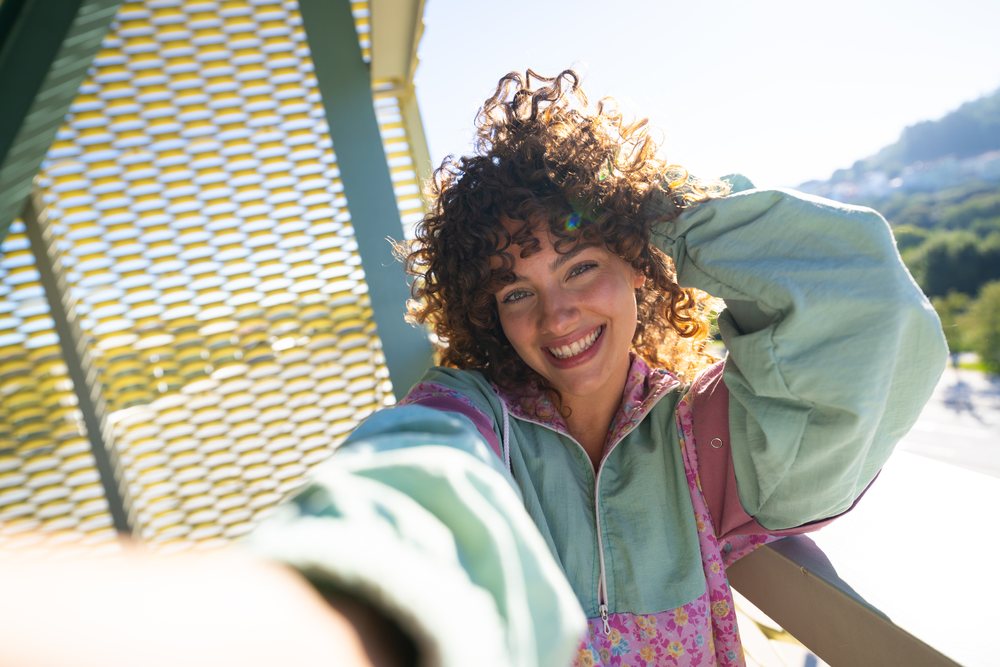 Woman with curly hair smiles taking a selfie outdoors near a modern architectural structure in Spain under sunlight.