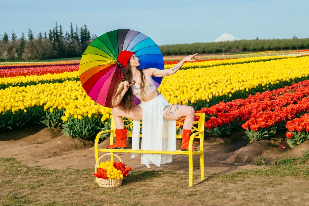 Woman with rainbow umbrella and red hat sitting on yellow bench in tulip field with mountain background, USA.