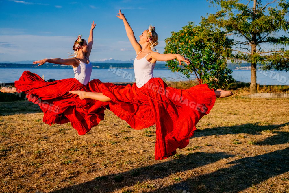 Two white women dancing and leaping outdoors in red skirts near water