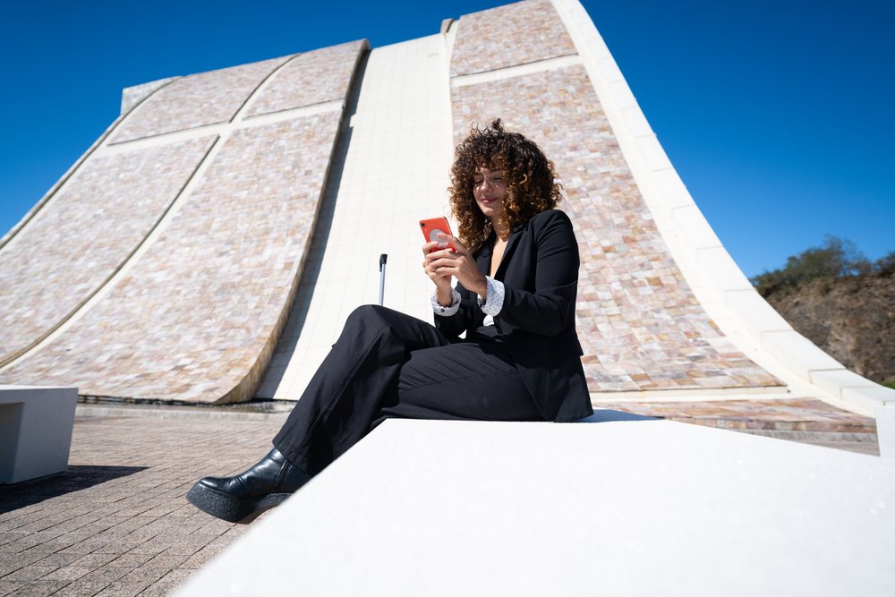 Businesswoman in black suit using smartphone while sitting outdoors near modern architecture, S, traveling executive, Spain, Santiago de Compostela.