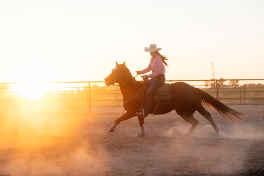 Girl wearing a cowboy hat rides a brown horse with western tack through a dusty outdoor arena at sunset, Southwest USA.