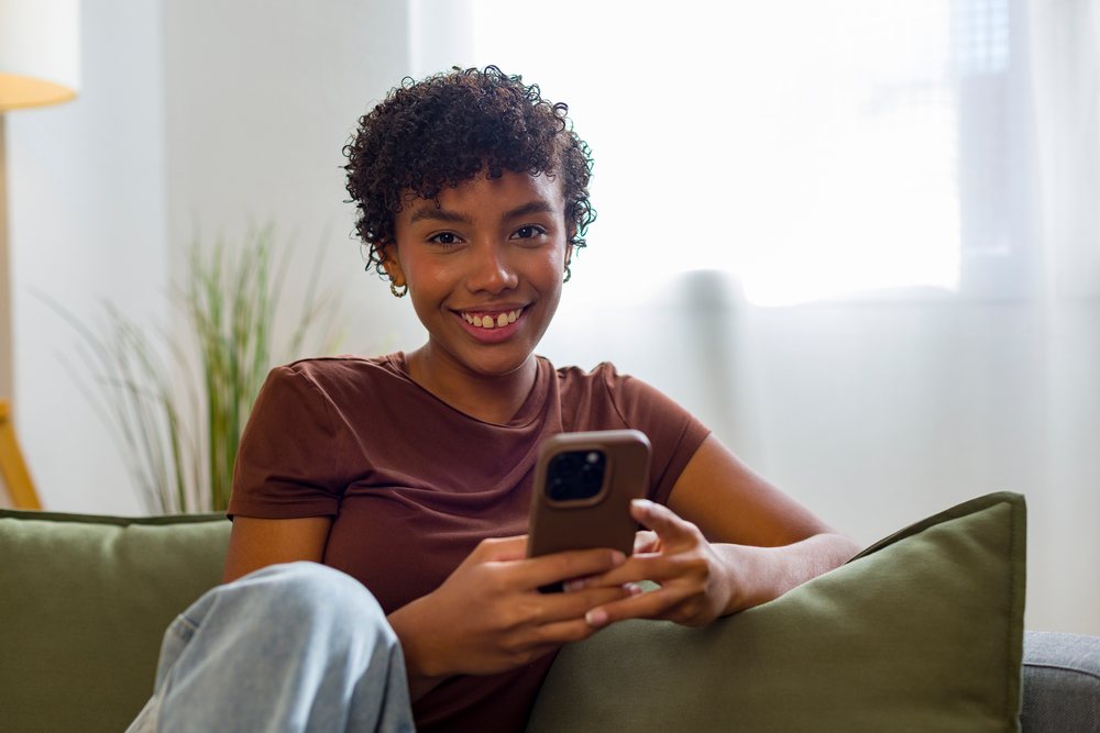 Young woman with curly hair smiling and using a smartphone while sitting on a couch with green cushions indoors at home.
