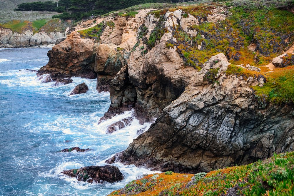 Rocky coastline with waves crashing against cliffs covered in green and orange vegetation on a California shore, USA.