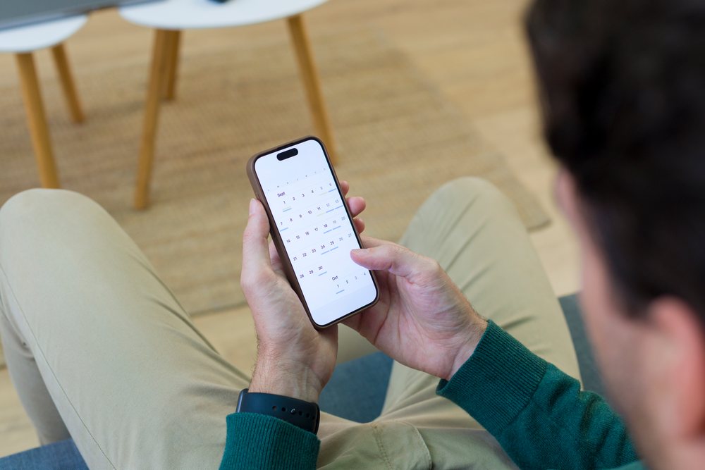 Person seated indoors holding a smartphone displaying a calendar app screen, Young adult, home.
