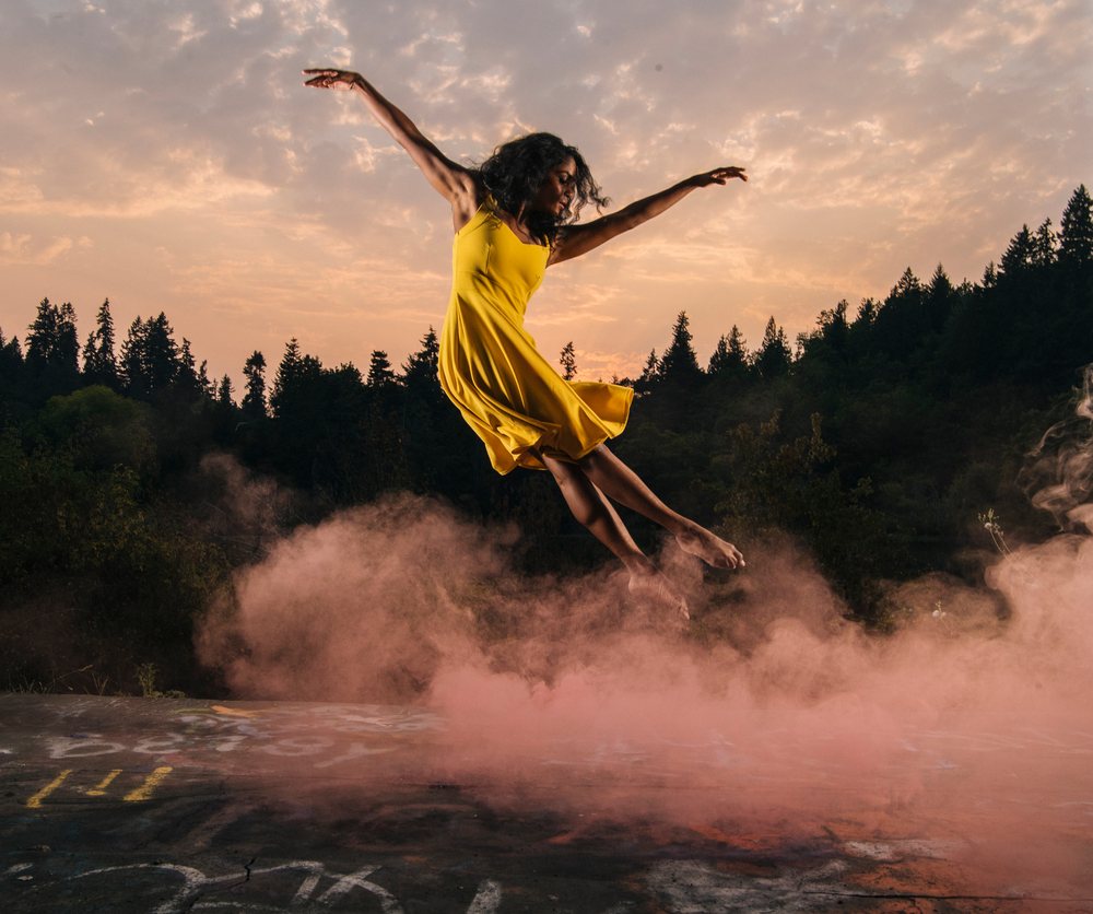 Barefoot woman in a yellow dress jumps outdoors among smoke and trees at sunset, USA.