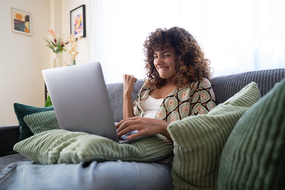 Woman with curly hair sits on a gray couch in a living room, cheering while using a laptop, home, girl, Spain.