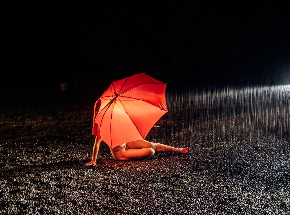 Woman sitting on wet pavement under a red umbrella during nighttime rain, cosplay, nudity, freedom, expression, USA.