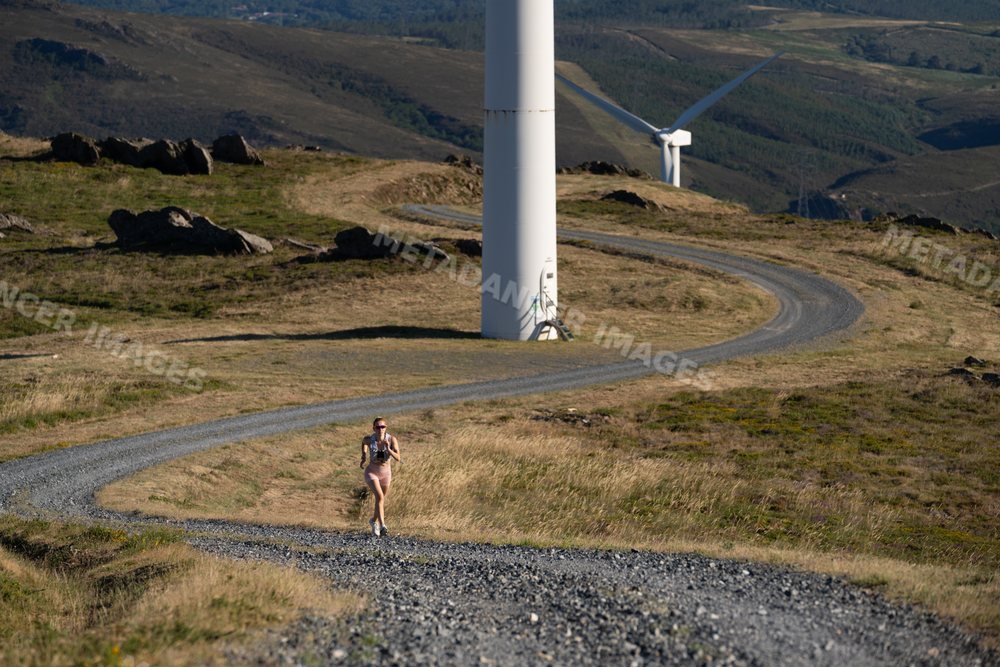 A white woman runs on a winding trail by wind turbines in Galicia