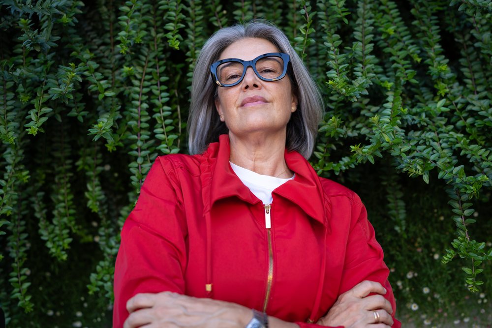 Confident middle-aged woman with gray hair and glasses stands outdoors against green leafy shrubbery, Spain.