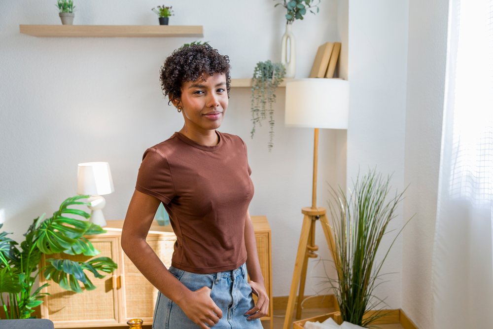 Young woman stands smiling in a cozy modern home interior with plants and natural window light.
