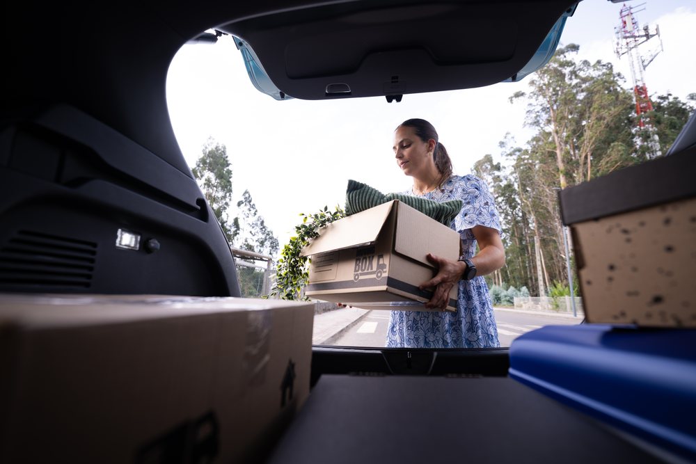 Woman loading a moving box containing a pillow and a green plant into the trunk of a parked car outdoors on a street with trees and a telecommunication tower, Spain.