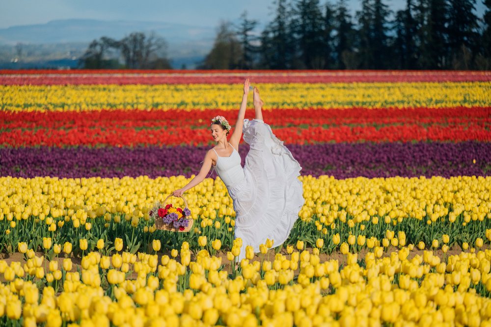 Ballet dancer in a white dress wearing a flower crown holds a basket of flowers while posing gracefully in a colorful tulip field, USA.
