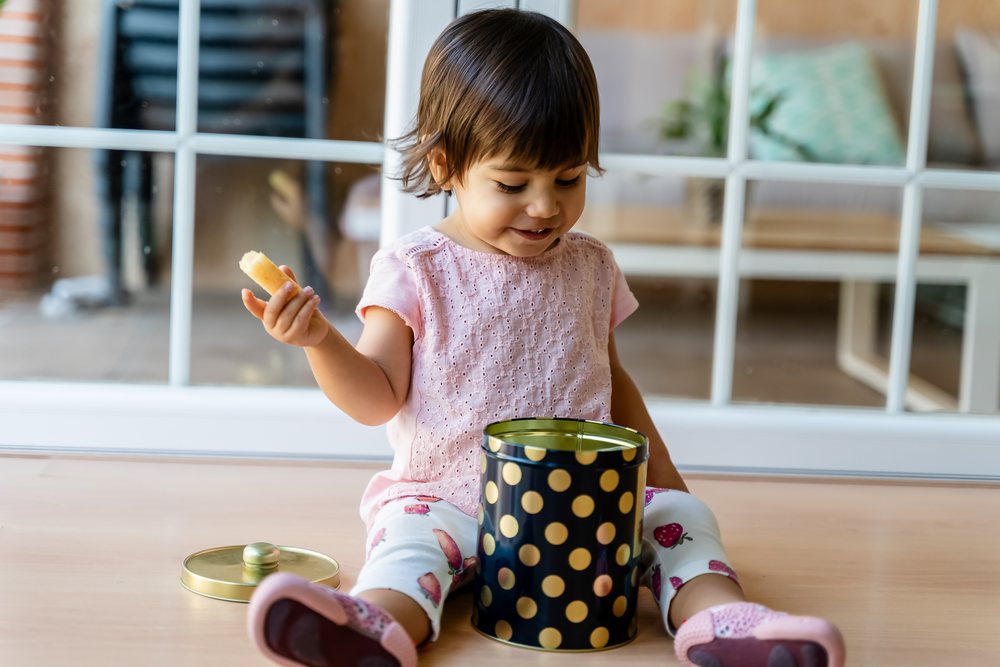 Toddler sitting on the floor indoors in Spain playing with a black cookie tin with gold polka dots and holding a cookie, kids.