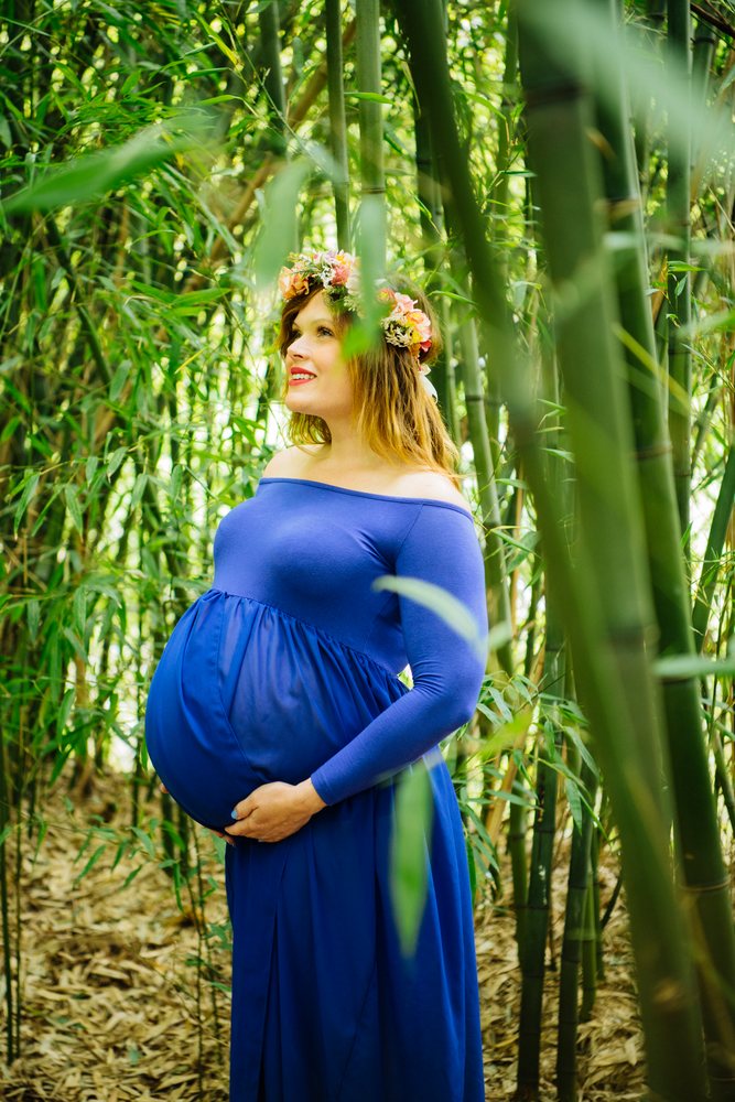 Pregnant woman wearing an off-shoulder blue dress and flower crown stands peacefully in a bamboo forest holding her belly, pregnancy.