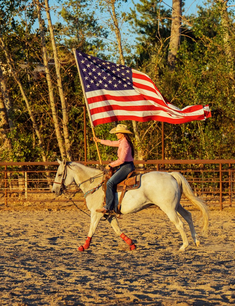 Woman riding white horse in fenced sandy outdoor arena holding upright American flag in Southwest USA daytime.