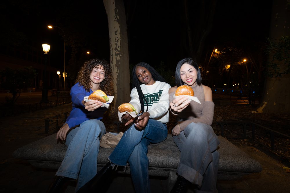 Three women sitting on a bench in an urban park at night offering hamburgers and smiling outdoors, Madrid, Spain.