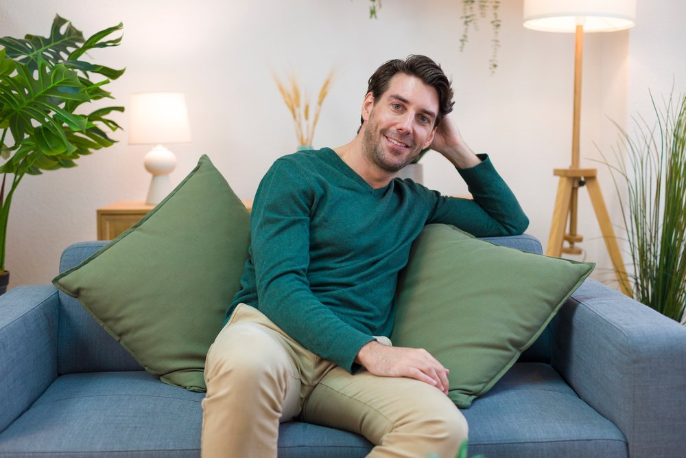 Man wearing green sweater and beige pants sits smiling on a sofa with green pillows in a modern living room at home, Technology, young adult.