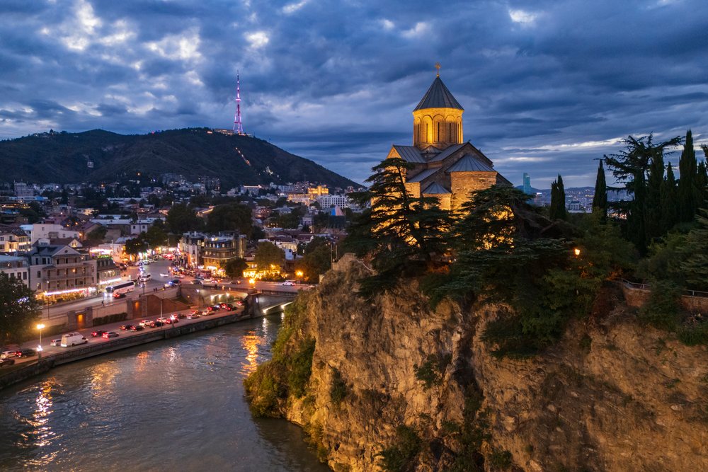 Metekhi Church illuminated on a cliff overlooks a river with Tbilisi cityscape and hills visible at Dusk, Georgia.