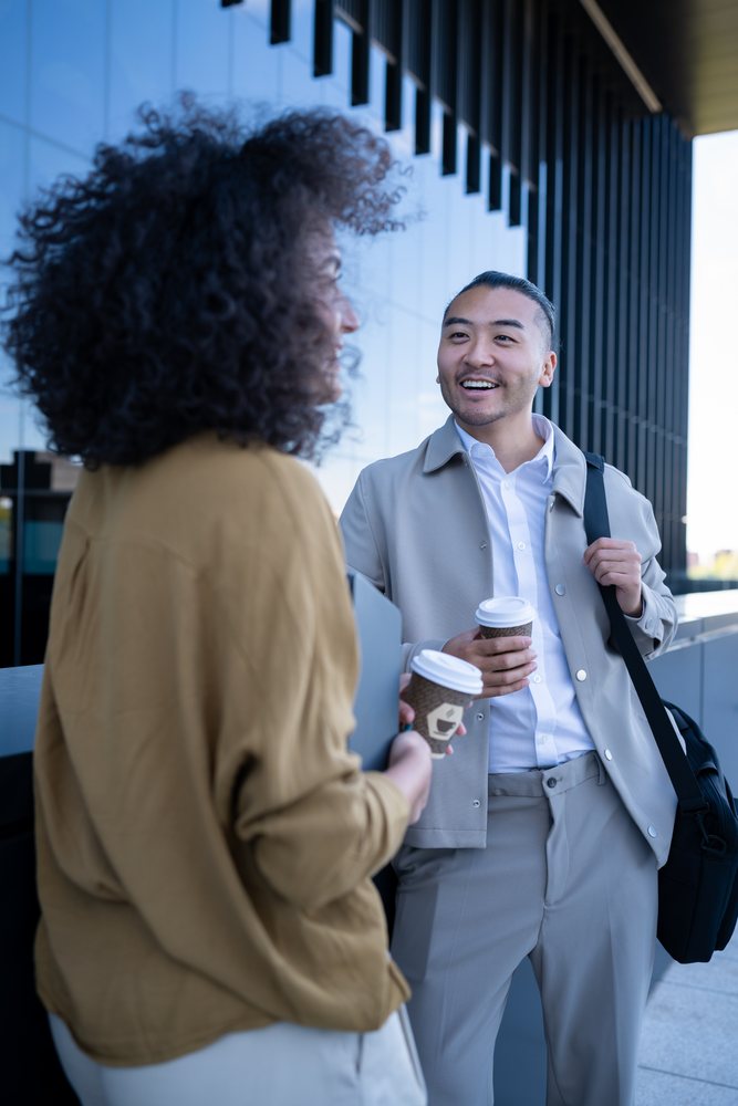 Two people holding disposable coffee cups talk and smile outside a modern office building in an urban sidewalk environment, Madrid, Spain.