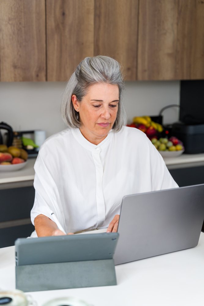 Middle aged woman using a laptop and writing on a tablet with a stylus in a modern home kitchen, Spain.