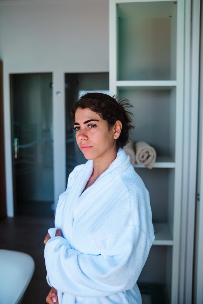 A woman wearing a white bathrobe stands indoors in a bathroom with shelving and towels visible in the, Maldives.
