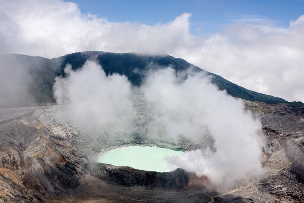 Steam rises over the pale green crater lake inside Poas Volcano crater under a blue sky.