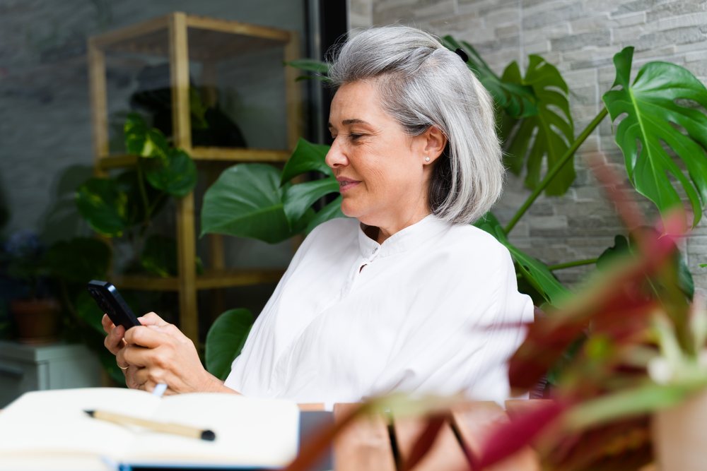 Woman with gray hair using a smartphone surrounded by indoor plants in a modern interior setting, Spain.
