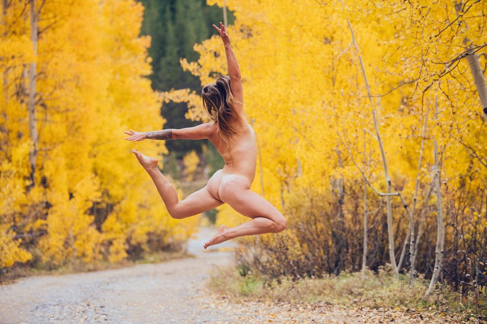 Nude woman jumping joyfully on a forest trail amid vibrant yellow autumn leaves, fall colors, Durango, Colorado, USA.