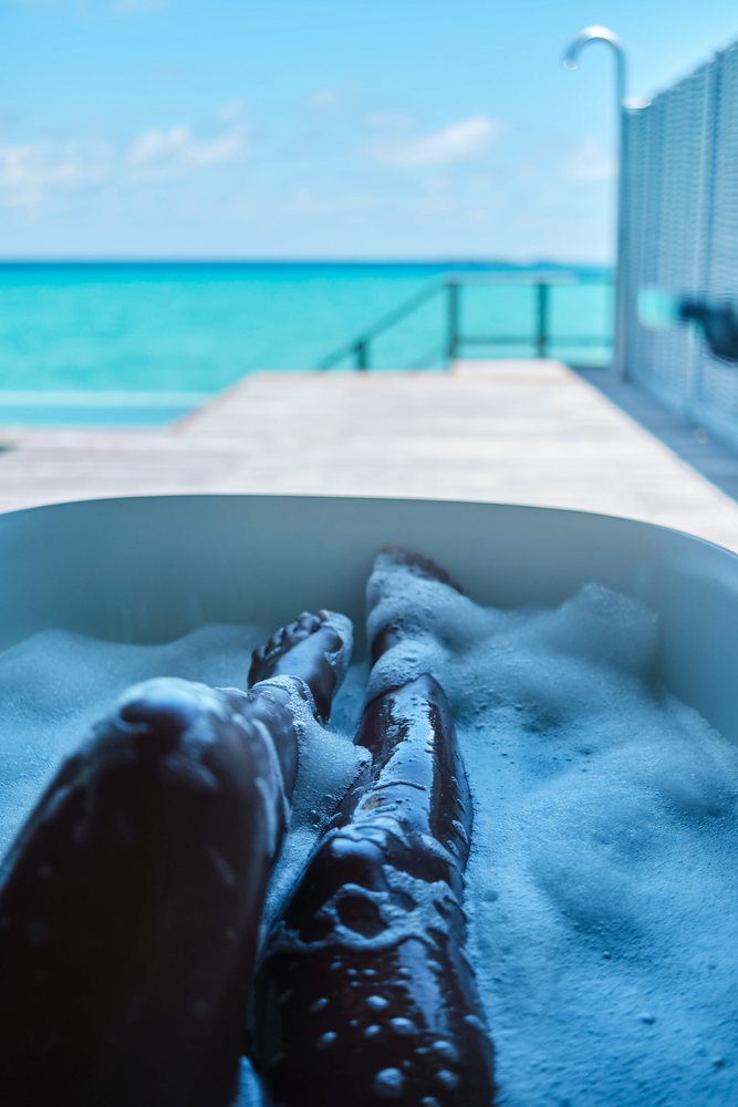 A person soaks their legs in a bubble bath with a view of the ocean and deck railing in the, Maldives.