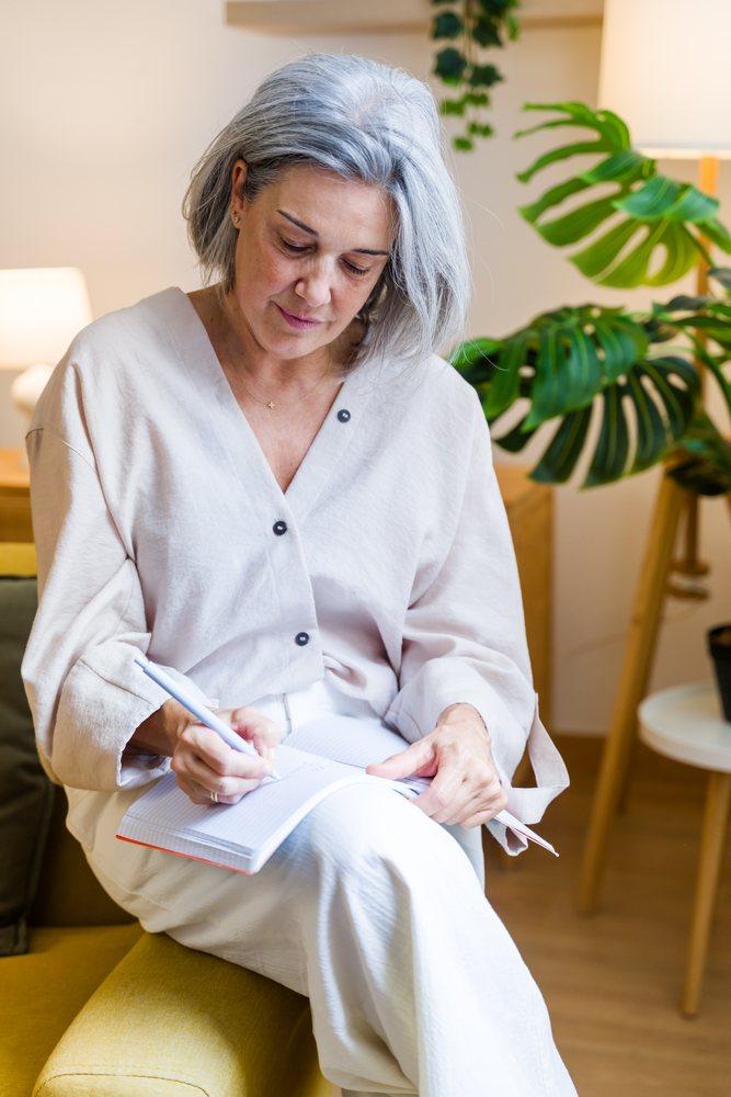 Middle-aged woman with gray hair writing in a notebook while seated on a yellow ottoman in a living room with indoor plants, Spain.
